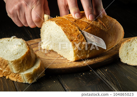 Slicing freshly baked wheat bread with a knife by the hands of a chef. National dish on the kitchen table 104865023