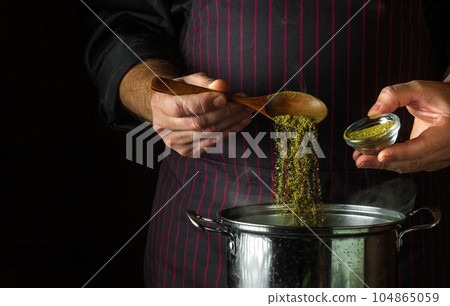 The cook adds dry spices to a pot of boiling food. Retsoran kitchen cooking concept with advertising space on black background. The cook adds dry spices to a pot of boiling food. Retsoran kitchen cooking concept with advertising space on black background. 104865059