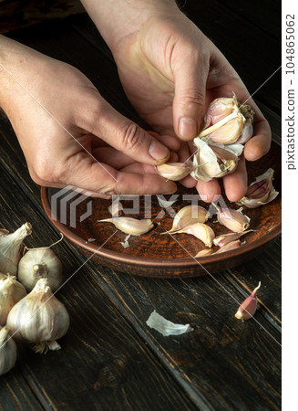 Close-up of the hands of a cook peeling garlic in the kitchen. The concept of cooking vegetarian food on a dark background Close-up of the hands of a cook peeling garlic in the kitchen. The concept of cooking vegetarian food on a dark background 104865062