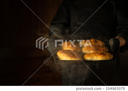 The chef holds a baking sheet with freshly baked buns or pies. Dark space for menu or recipe. Presentation of delicious pies. The chef holds a baking sheet with freshly baked buns or pies. Dark space for menu or recipe. Presentation of delicious pies. 104865070