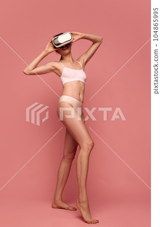 Full-length portrait of young woman with slim body, wearing Vr glasses, training in underwear against pink studio background 104865995