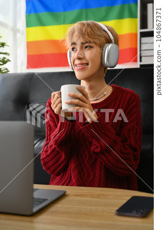 Satisfied young gay man in knitted sweater enjoy morning coffee and listening to music on wireless headphones Satisfied young gay man in knitted sweater enjoy morning coffee and listening to music on wireless headphones 104867371