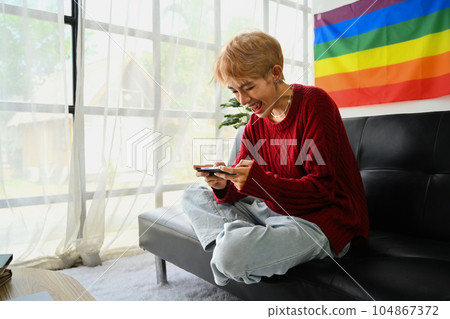 Cheerful gay man playing online game application on mobile phone while sitting in living room with rainbow flag 104867372
