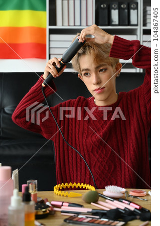 Attractive gay man in red sweater straightening hair with flat iron. LGBTQ, domestic life, fashion and beauty concept 104867405