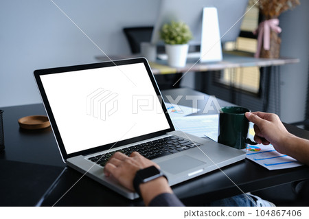 Closeup young businessman holding coffee cup and using laptop computer on black office desk 104867406