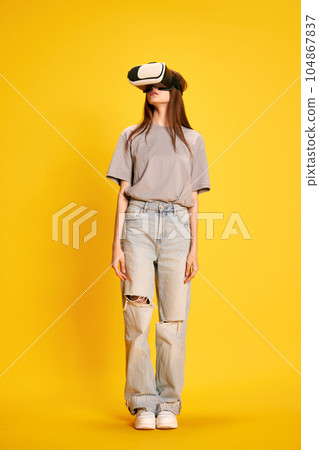 Full-length portrait of young beautiful girl in casual clothes, wearing vr glasses against yellow studio background 104867837