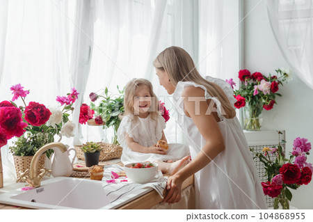 A little blonde girl with her mom on a kitchen countertop decorated with peonies. The concept of the relationship between mother and daughter. Spring atmosphere. A little blonde girl with her mom on a kitchen countertop decorated with peonies. The concept of the relationship between mother and daughter. Spring atmosphere. 104868955
