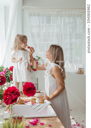 A little blonde girl with her mom on a kitchen countertop decorated with peonies. The concept of the relationship between mother and daughter. Spring atmosphere. A little blonde girl with her mom on a kitchen countertop decorated with peonies. The concept of the relationship between mother and daughter. Spring atmosphere. 104868962