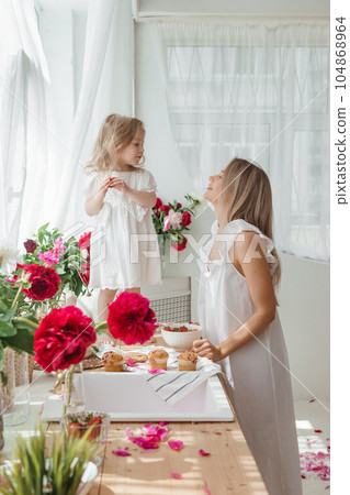 A little blonde girl with her mom on a kitchen countertop decorated with peonies. The concept of the relationship between mother and daughter. Spring atmosphere. A little blonde girl with her mom on a kitchen countertop decorated with peonies. The concept of the relationship between mother and daughter. Spring atmosphere. 104868964