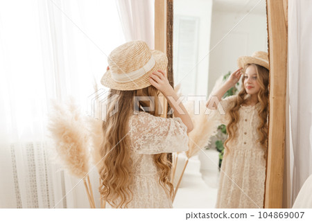 A beautiful teenage girl with long hair measures a straw hat in front of a mirror. Self-admiration of a blonde. A beautiful teenage girl with long hair measures a straw hat in front of a mirror. Self-admiration of a blonde. 104869007