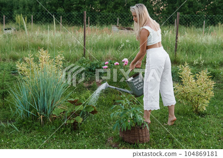 A blonde woman is watering flowers in the garden in the courtyard of a village house. 104869181