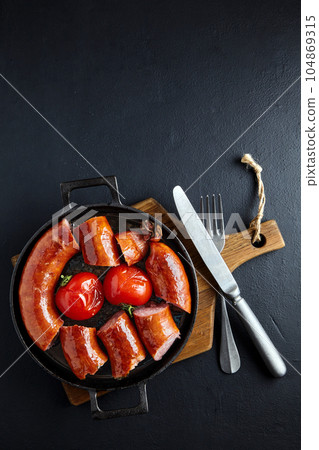 sliced fried pork sausage in a cast iron pan with tomatoes and cutlery on a wooden cutting board on a dark concrete table sliced fried pork sausage in a cast iron pan with tomatoes and cutlery on a wooden cutting board on a dark concrete table 104869315