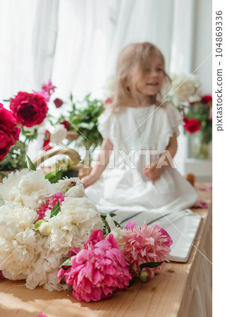 A little blonde girl on a kitchen countertop decorated with peonies. Spring atmosphere. A little blonde girl on a kitchen countertop decorated with peonies. Spring atmosphere. 104869336
