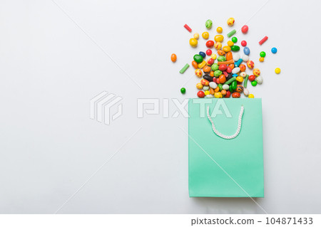 shopping paper gift bag in corner full of assorted traditional candies falling out on colored background with copy space. Happy Holidays sale concept 104871433