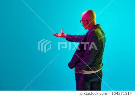 Side view portrait of young bald man in casual clothes showing away against blue studio background in neon light 104871514
