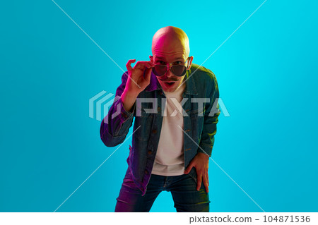 Portrait of young bald man with moustache wearing stylish clothes and sunglasses against blue studio background in neon light Portrait of young bald man with moustache wearing stylish clothes and sunglasses against blue studio background in neon light 104871536
