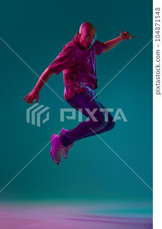 Full-length portrait of young bald man with moustache in stylish clothes emotionally jumping against cyan studio background in neon light 104871548