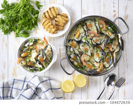 Mussels clams in cream sauce in cooking pan and toasted baguet on wooden table, top view. 104872639