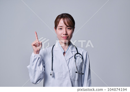 A young female doctor in a white coat who raises her index finger A young female doctor in a white coat who raises her index finger 104874194