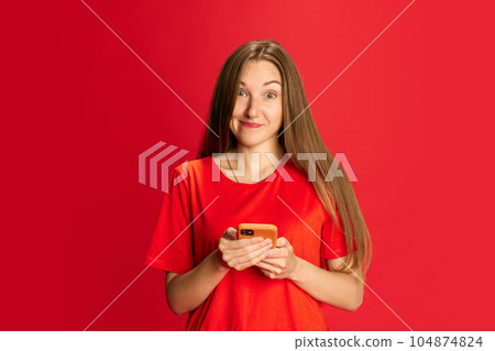 Portrait of beautiful young woman with joyful face texting messages on mobile phone against red studio background. Communication 104874824