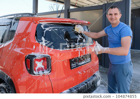 Happy man washes a car at a car wash. Car in foam Happy man washes a car at a car wash. Car in foam 104874855