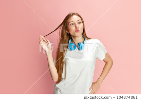 Portrait of young beautiful girl in casual clothes and headphones posing with grimacing funny face against pink studio background. 104874861