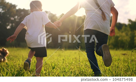 children and dog run. a boy and a girl holding hands run through the grass in the summer at sunset in the park. happy family kid dream concept. children run at sunset in summer park sunlight children and dog run. a boy and a girl holding hands run through the grass in the summer at sunset in the park. happy family kid dream concept. children run at sunset in summer park sunlight 104875498