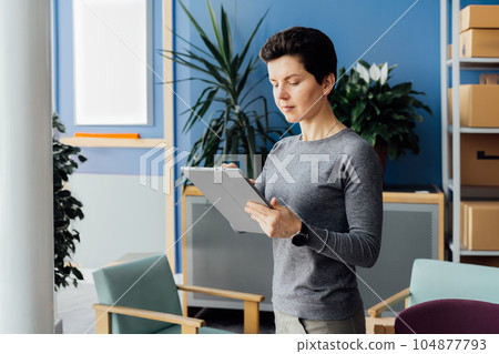 Neutral gender middle-aged businesswoman making notes on tablet, checking information before sending to customer. Entrepreneur small business. Orders Ready for Shipment. Logistic, delivery. 104877793