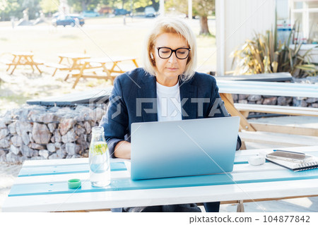 Concentrated 50's stylish, confident mature business woman, middle aged lady using wireless ear buds and laptop, working remotely in park. Caucasian woman make a video call, study, work online lesson. Concentrated 50's stylish, confident mature business woman, middle aged lady using wireless ear buds and laptop, working remotely in park. Caucasian woman make a video call, study, work online lesson. 104877842