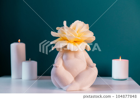 Close up white table with ceramic female body-shaped vase with rose flower, burning candles on turquoise blue wall background. Zen, cozy, meditation female bedroom decor. Self recovery place at home. Close up white table with ceramic female body-shaped vase with rose flower, burning candles on turquoise blue wall background. Zen, cozy, meditation female bedroom decor. Self recovery place at home. 104877848