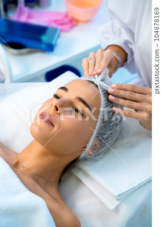 Woman laying in cosmetology clinic in disposable hat preparing for skin care beautician procedures. Woman laying in cosmetology clinic in disposable hat preparing for skin care beautician procedures. 104878169