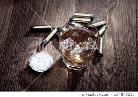 Whisky shot with ice and ammunitions with old silver dollars coins on wooden table 104878285