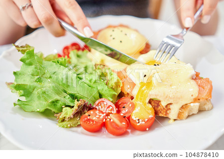 Woman cuts poached egg and yolk flows out. Brioche, avocado cream with salmon and egg . Healthy breakfast, protein. Restaurant dish. Woman cuts poached egg and yolk flows out. Brioche, avocado cream with salmon and egg . Healthy breakfast, protein. Restaurant dish. 104878410