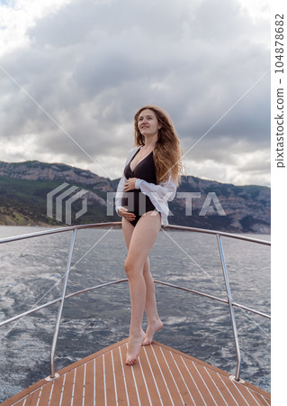 Woman on a yacht. Happy model in a swimsuit posing on a yacht against a blue sky with clouds and mountains Woman on a yacht. Happy model in a swimsuit posing on a yacht against a blue sky with clouds and mountains 104878682