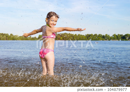 Happy child plays with splashes of water on the river bank 104879977