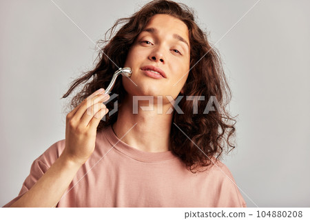 Young handsome long haired guy using de-puffing face roller to keep his skin soft and smooth. Caucasian man with brown hair maintaining his natural beauty with skincare routine. Men's self-care. Young handsome long haired guy using de-puffing face roller to keep his skin soft and smooth. Caucasian man with brown hair maintaining his natural beauty with skincare routine. Men's self-care. 104880208