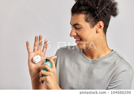 Happy young smiling guy putting facial wash on his fingers, preparing to apply the beauty product on his face. Swarthy man with black curly hair uses hydrating skin cleanser in his skincare routine. Happy young smiling guy putting facial wash on his fingers, preparing to apply the beauty product on his face. Swarthy man with black curly hair uses hydrating skin cleanser in his skincare routine. 104880209