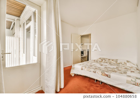 a bedroom with red carpet and white walls, there is a small bed in the room next to the window 104880658