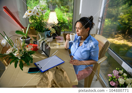 Latin American pregnant woman taking sip of coffee, sitting at table and working on clipboard in a floral design studio 104880771