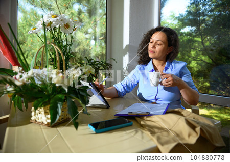 Charming woman floral designer holding a cup of coffee, sketching on digital tablet, working on new flower arrangements 104880778