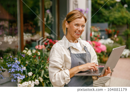 Small flower shop owner florist wearing apron working on laptop and taking online orders in store Small flower shop owner florist wearing apron working on laptop and taking online orders in store 104881179