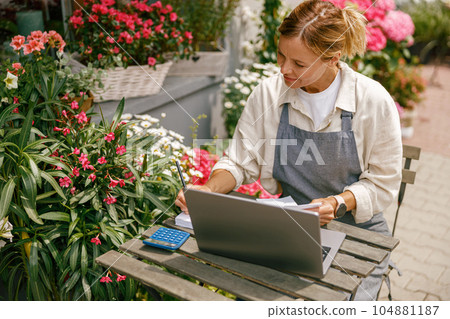 Professional flower shop owner florist using laptop and writing on notebook at flower shop Professional flower shop owner florist using laptop and writing on notebook at flower shop 104881187