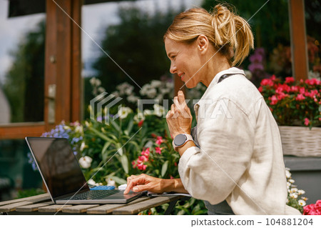 Small flower shop owner florist wearing apron working on laptop and taking online orders in store 104881204