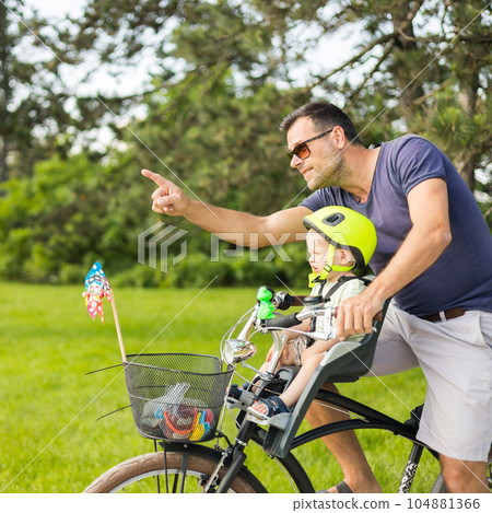 Look over there. Active family day in nature. Father and son ride bike through city park on unny summer day. A cute boy is sitting in front bicycle chair while father rides bicycle. Father son bonding Look over there. Active family day in nature. Father and son ride bike through city park on unny summer day. A cute boy is sitting in front bicycle chair while father rides bicycle. Father son bonding 104881366