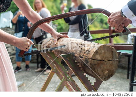 Young bridal couple groom bride sawing a tree trunk together german wedding tradition Young bridal couple groom bride sawing a tree trunk together german wedding tradition 104882568