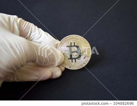 a hand in a glove holds a gold-colored bitcoin coin on a black background 104882696