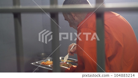 Elderly criminal in orange uniform sits in prison cell, eats dinner 104883214