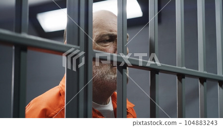 Face close up of elderly prisoner standing behind metal bars in prison cell 104883243