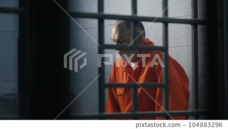 Criminal in orange uniform sits on prison bed Criminal in orange uniform sits on prison bed 104883296