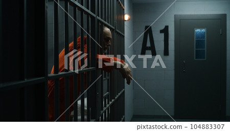 Upset inmate stands behind bars in jail or correctional facility Upset inmate stands behind bars in jail or correctional facility 104883307